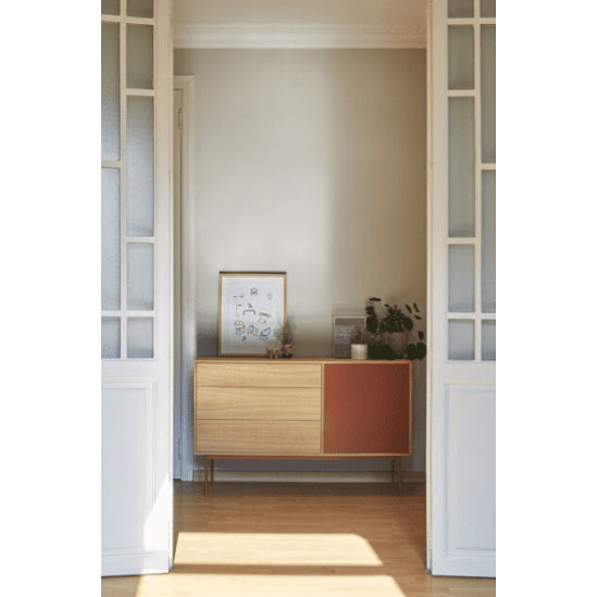 En una habitación minimalista, el aparador moderno pequeño Yoko 1P3C con detalles en rojo ofrece un almacenamiento eficiente para un cuadro enmarcado, una vela y una planta. Se encuentra entre puertas dobles blancas sobre un piso de madera, lo que encarna el diseño moderno.