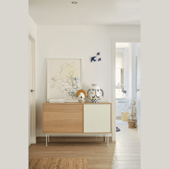 El aparador moderno pequeño Yoko 1P3C ejemplifica el almacenamiento eficiente con una pieza de arte abstracto enmarcada y dos jarrones decorativos en la parte superior, ubicados en una habitación luminosa con piso de madera clara.