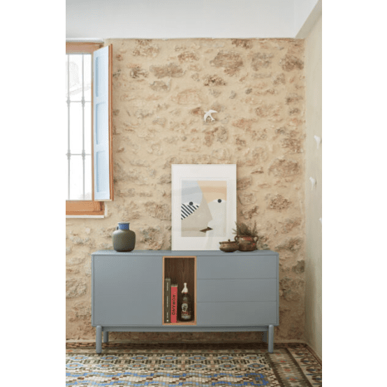 Un interior con pared de piedra resalta el Aparador pequeño con cajones Corvo 1P3C, adornado con libros y decoración de diseño escandinavo. Una obra de arte abstracta enmarcada realza el espacio, mientras la luz se cuela a través de una ventana abierta sobre la alfombra estampada que se encuentra debajo.