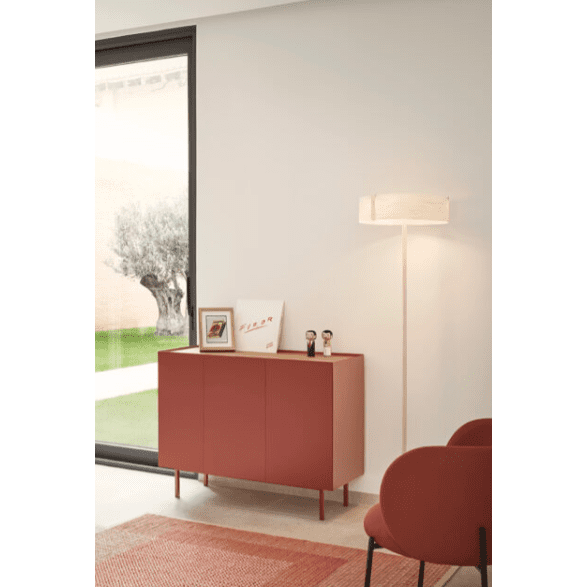 Una habitación contemporánea muestra un Aparador pequeño con Puertas Arista 3P3C en rojo intenso con dos figuras y obras de arte enmarcadas. Una elegante lámpara de pie se encuentra al lado, mientras que una gran ventana ofrece una vista al jardín, lo que realza el diseño contemporáneo.