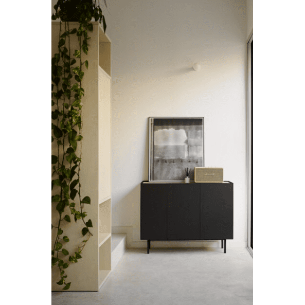 El interior minimalista cuenta con un Aparador pequeño con Puertas Arista 3P3C para almacenamiento elegante, complementado con obras de arte abstractas y un letrero decorativo. A la izquierda, una estantería alta con plantas colgantes descansa contra una pared de color claro, lo que realza el diseño contemporáneo.
