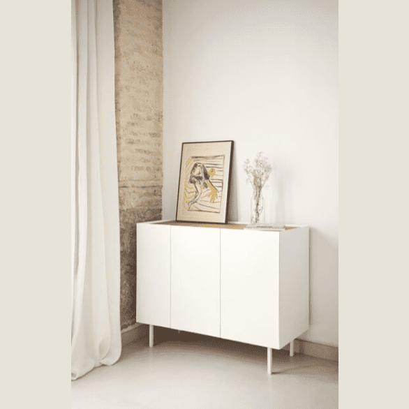 El Aparador pequeño con Puertas Arista 3P3C, un elegante armario blanco, se encuentra contra la pared adornado con un cuadro y un jarrón de flores secas. Una cortina cae suavemente a su izquierda, lo que le aporta elegancia a esta pieza de almacenaje compacta.