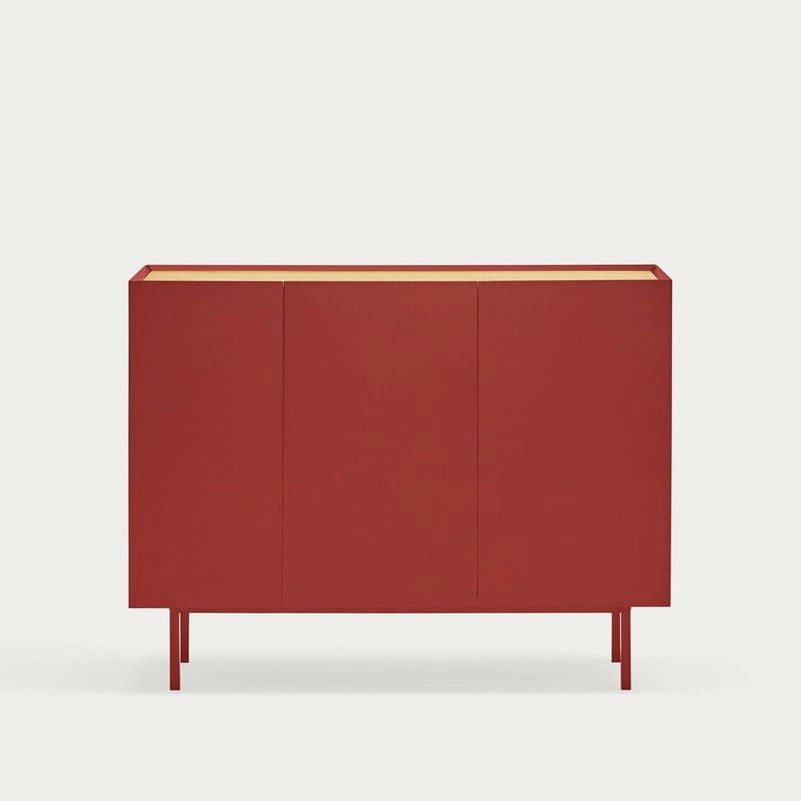 El Aparador pequeño con Puertas Arista 3P3C es un armario minimalista rojo con tres puertas, elevado sobre patas delgadas, que muestra un elegante almacenamiento con su diseño contemporáneo sobre un fondo blanco liso.