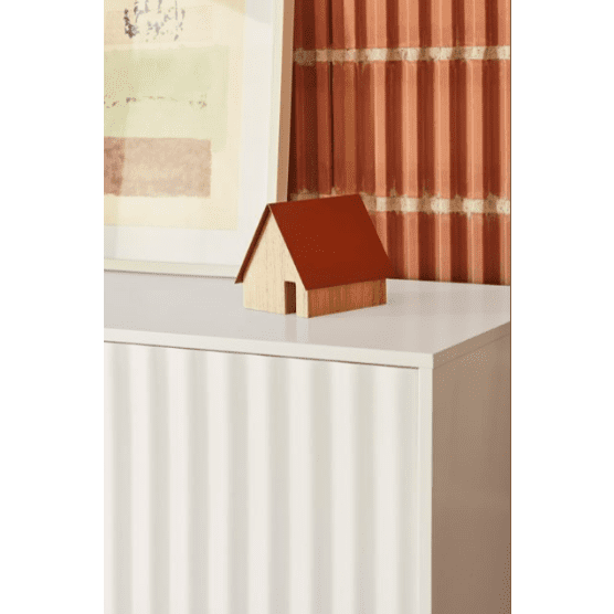 El Aparador pequeño con Puertas Sierra 3P3C, con un techo rojo y un diseño en forma de casa, se encuentra sobre una superficie blanca sobre un fondo de patrones de ladrillos y arte abstracto, que encarna el estilo industrial moderno.