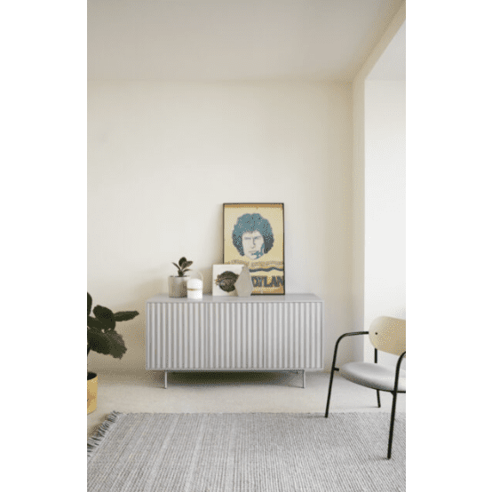 Una habitación minimalista cuenta con un armario de MDF lacado gris con amplio espacio de almacenamiento, adornado con un retrato enmarcado, plantas y una vela. Un Aparador pequeño con Puertas Sierra 3P3C agrega carácter mientras que una silla moderna y una alfombra gris completan la escena serena en primer plano.