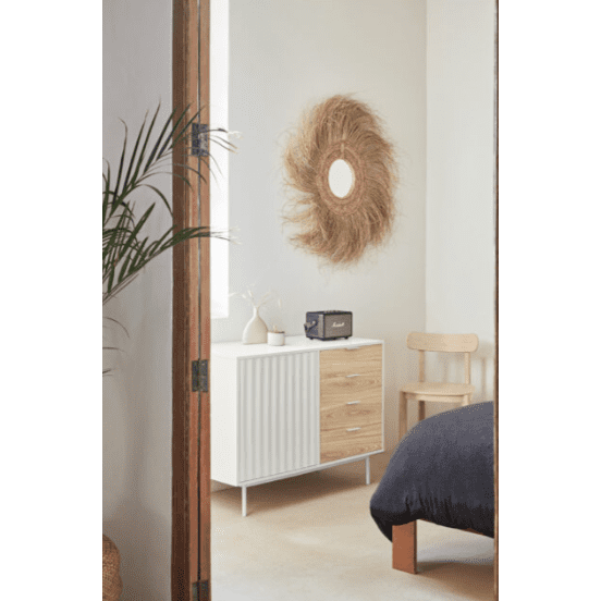 Acogedor dormitorio con un Aparador pequeño en Madera y DM Sierra 1P4C, complementado con un espejo con marco de paja, un altavoz negro y un florero. Una silla de madera aporta calidez mientras que la manta oscura de la cama parcialmente visible realza el diseño moderno de la habitación.