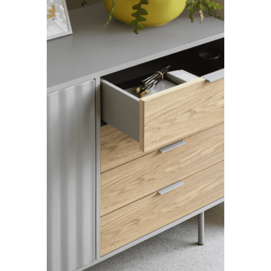 El Aparador pequeño en Madera y DM Sierra 1P4C, con acabado en gris y madera, incluye un cajón para utensilios abierto. Su diseño asimétrico muestra elegantemente una planta y un marco de fotos en la parte superior, aportando estilo e inteligentes soluciones de almacenaje.