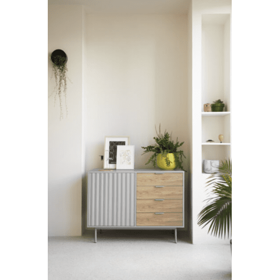 El Aparador pequeño en Madera y DM Sierra 1P4C exhibe elegantemente obras de arte enmarcadas y plantas con sus puertas grises y cajones de madera. Este aparador moderno ofrece soluciones de almacenaje y un diseño asimétrico, y se destaca elegantemente contra una pared clara junto a un estante incorporado.