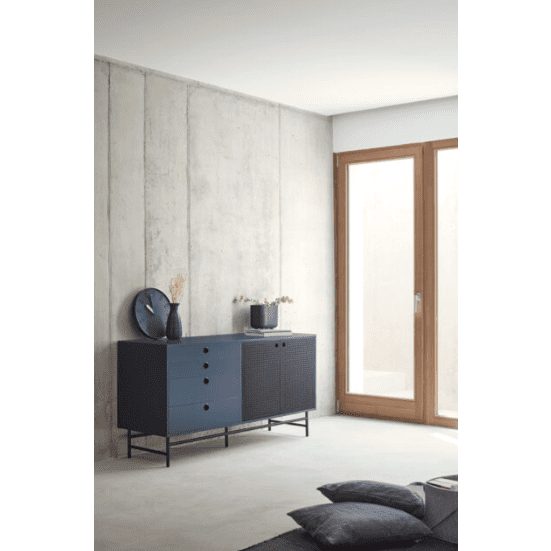 Una habitación minimalista resalta el estilo industrial con un aparador azul contra una pared de hormigón claro. Un elegante reloj redondo y un jarrón adornan el Aparador pequeño en Metal y DM Punto 2P4C, mientras que cerca, unos cojines se encuentran junto a una gran puerta de vidrio con marco de madera.