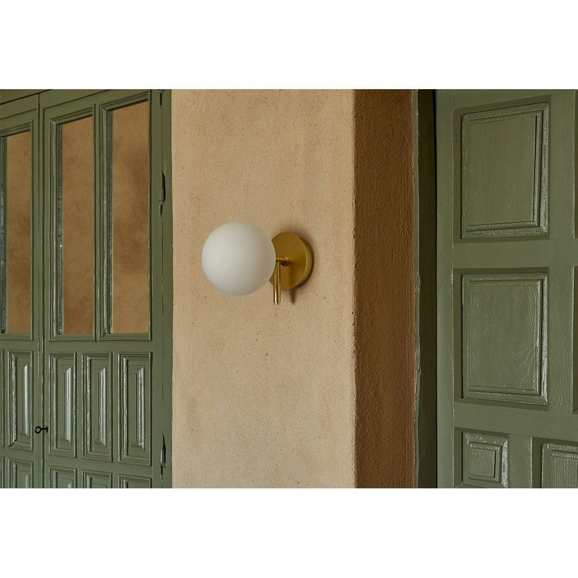 El aplique de pared Atom, una luminaria esférica de pared con base de latón sobre una pared beige, personifica la iluminación minimalista junto a puertas y ventanas con paneles verdes.