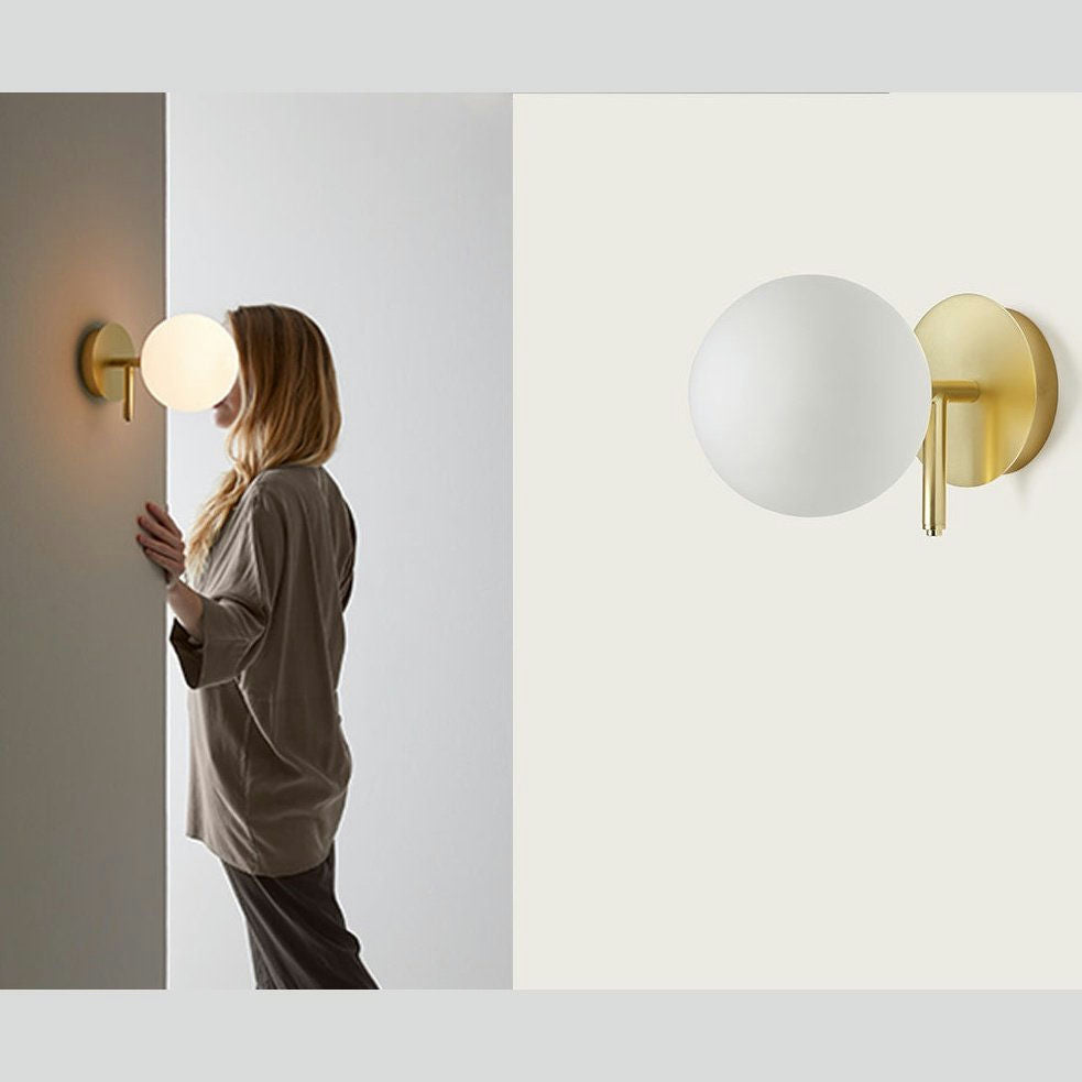 Una persona se encuentra frente a una pared adornada con el aplique de pared Atom, que muestra su diseño moderno. La lámpara esférica también se muestra por separado a la derecha sobre un fondo claro, lo que resalta su estilo de iluminación elegante y minimalista.