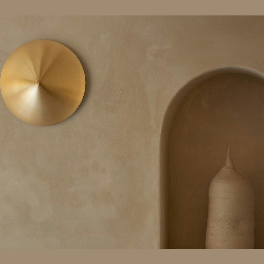 Una pared beige minimalista presenta una escultura cónica abstracta en una alcoba, complementada por una decoración de pared metálica circular y realzada por el elegante Aplique de pared Hat.