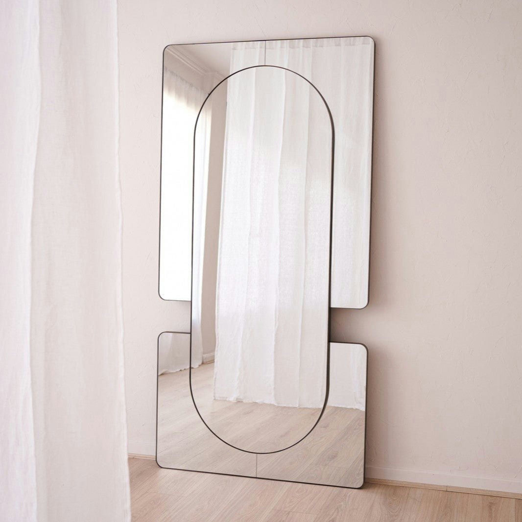 El Espejo de Suelo en Metal con Acabado Envejecido N2, alto y rectangular, destaca con su diseño excepcional al apoyarse contra una pared clara junto a cortinas translúcidas sobre un suelo de madera.