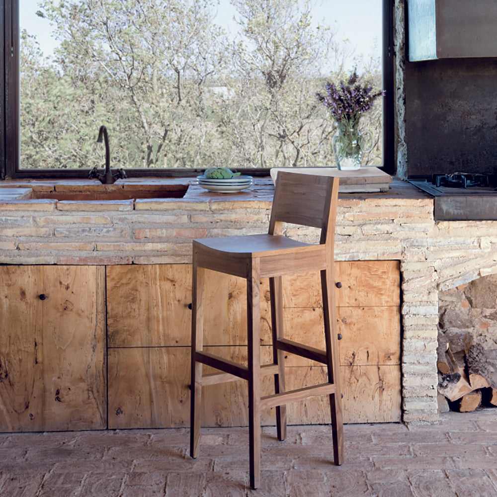 El Taburete alto en madera teca Andrea aporta un diseño moderno a la cocina rústica con sus elementos de ladrillo y madera, complementando el encanto natural del espacio cerca de una gran ventana y un fregadero de piedra.