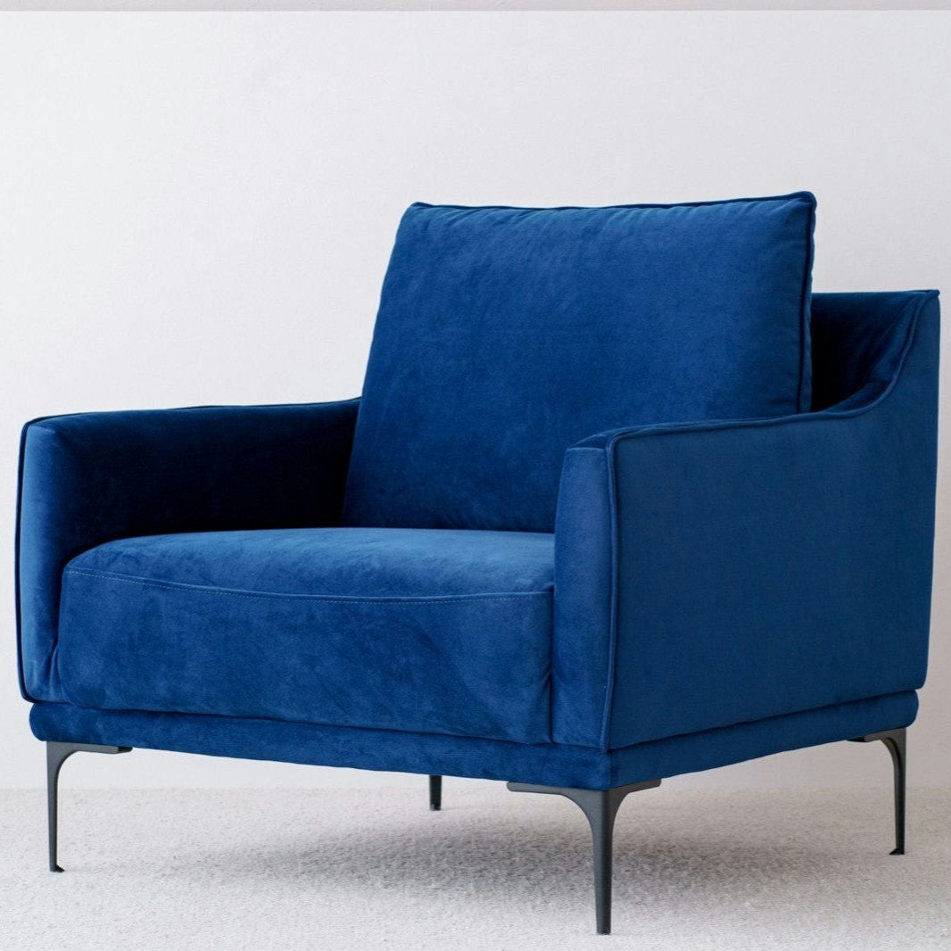 La Butaca en Terciopelo Azul Noche Helia, con elegantes patas de metal, personifica el estilo de interiorismo. Ubicado lujosamente sobre una alfombra clara, rezuma lujo y comodidad.