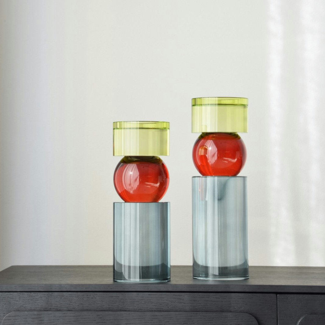 Dos candelabros Cristal Color Cilindro descansan sobre una superficie de madera oscura, con tapas verdes, centros redondeados rojos y bases cilíndricas. Su diseño geométrico moderno realza los interiores contemporáneos con estilo escultural moderno.