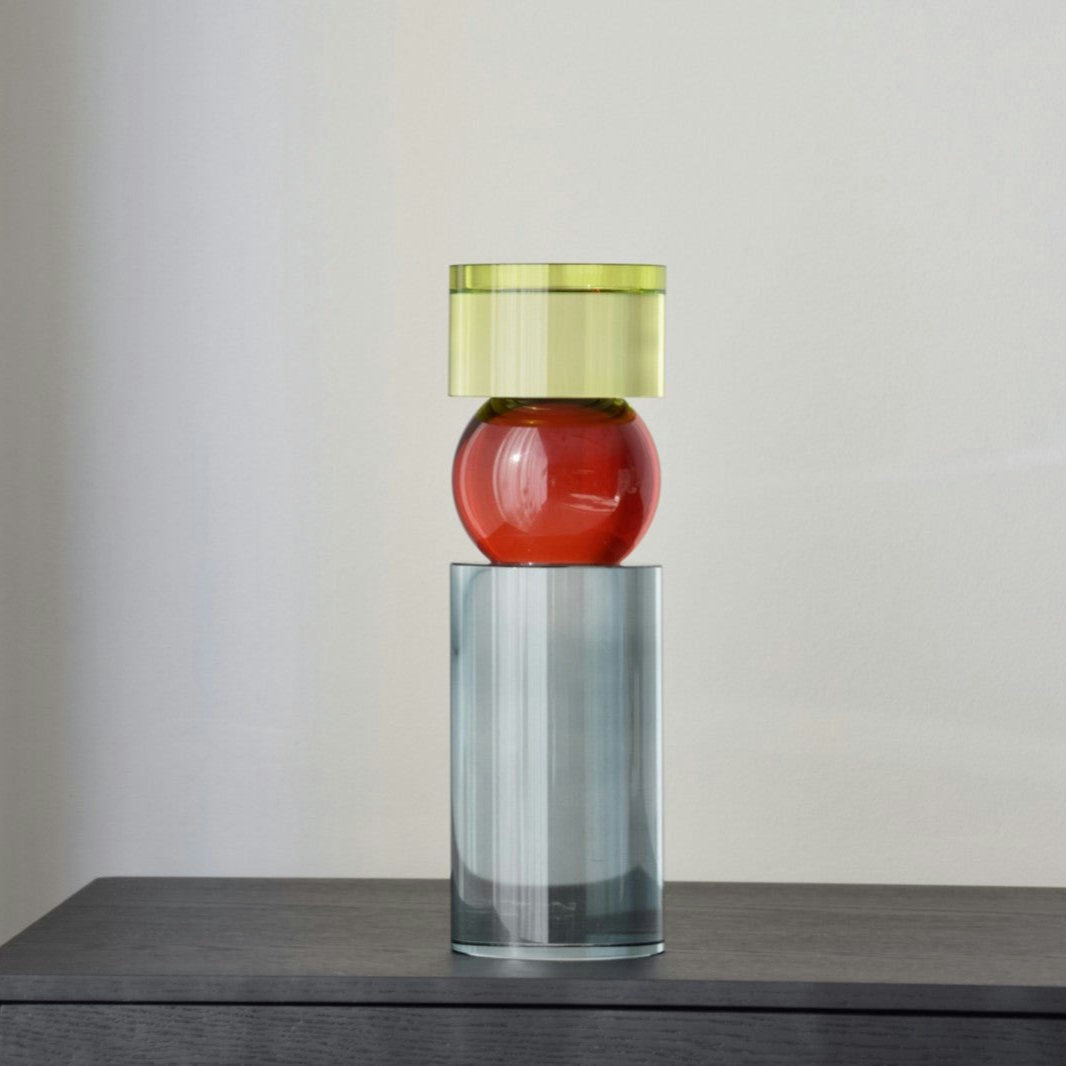 El Candelabros Cristal Color Cilindro (Grande y Pequeño) presenta una base cilíndrica transparente, un centro esférico rojo y una tapa circular verde sobre una superficie oscura contra una pared lisa, resaltando sus colores vibrantes en diseño escultural y moderno.