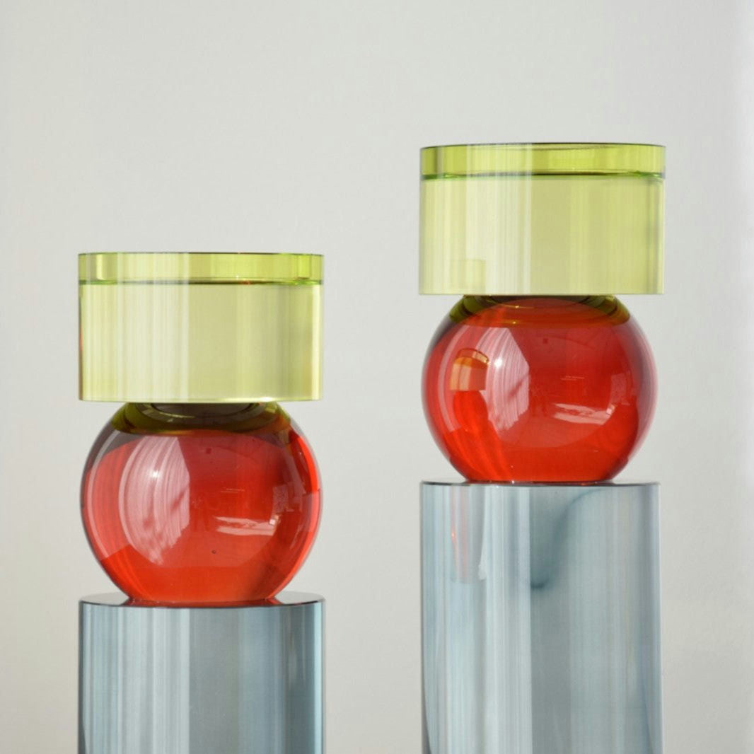 Los candelabros Cristal Color Cilindro, tanto el grande como el pequeño, presentan un diseño escultural y moderno: tapas cilíndricos verdes sobre bases esféricas rojas, situados en pilares azul-gris frente a un fondo neutro.