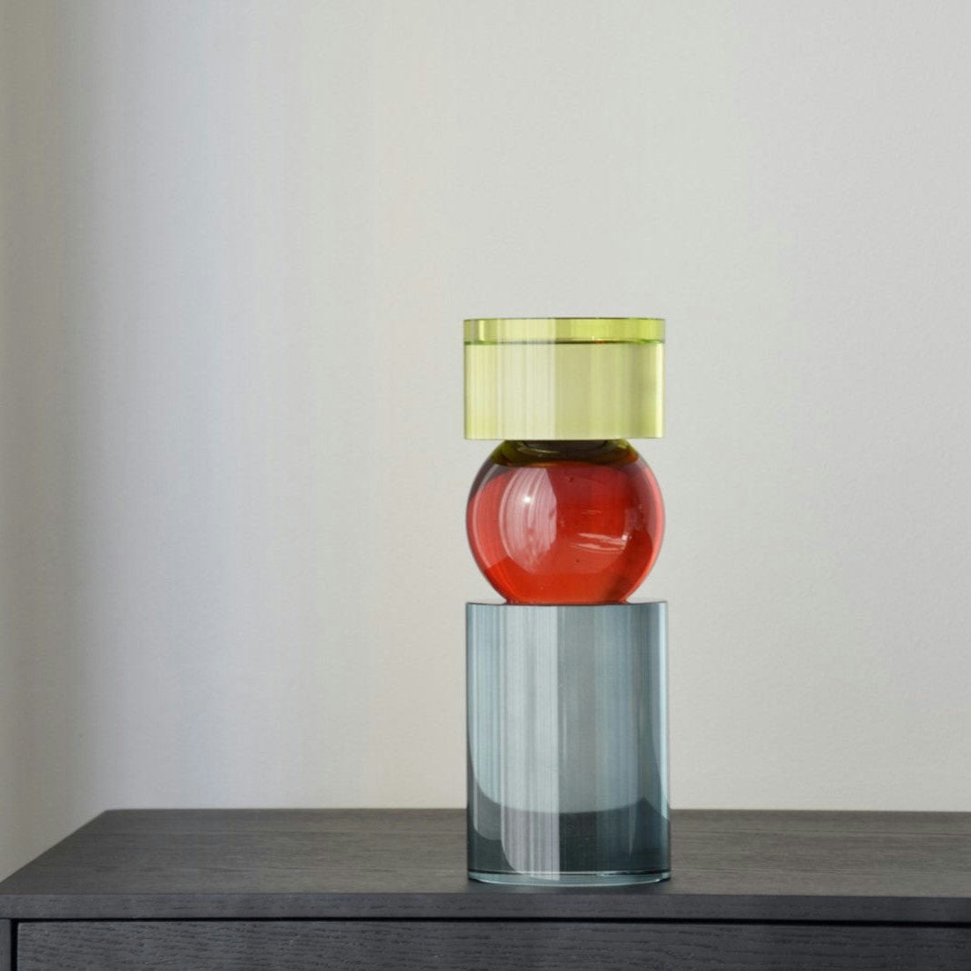 El Candelabros Cristal Color Cilindro presenta esculturas de vidrio vibrantes en tres formas geométricas: una parte superior cilíndrica verde, un centro esférico rojo y una base cilíndrica gris, elegantemente colocadas sobre una superficie oscura contra una pared blanca lisa.