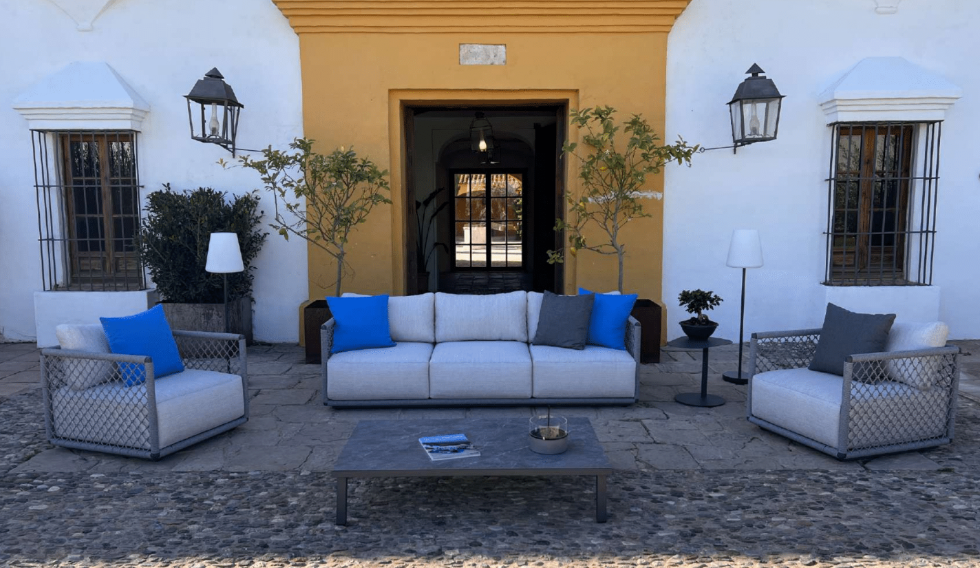 Conjunto de patio exterior con un Sofá 3 plazas de exterior OCEAN (aluminio y cuerda gris, 220×83×62 cm, estilo moderno), dos sillones con cojines azules y grises, mesa de centro y plantas en macetas frente a un edificio blanco y amarillo.