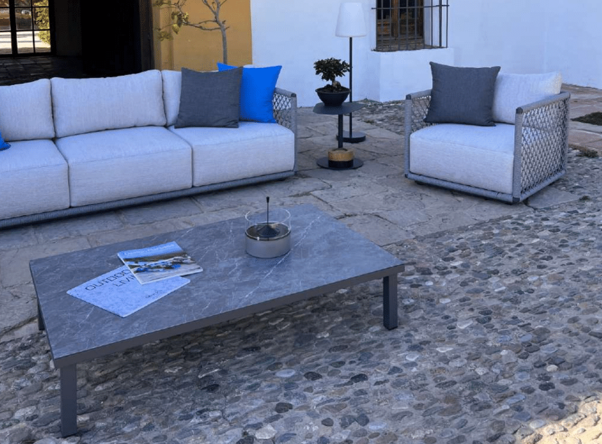 Moderno patio exterior con muebles de jardín modernos, con sofá gris, sillón a juego, cojines azules y grises, y la mesa de centro de exterior OCEAN de aluminio y cristal cerámico (125×75×28 cm) sobre una superficie de adoquines y losas de piedra.