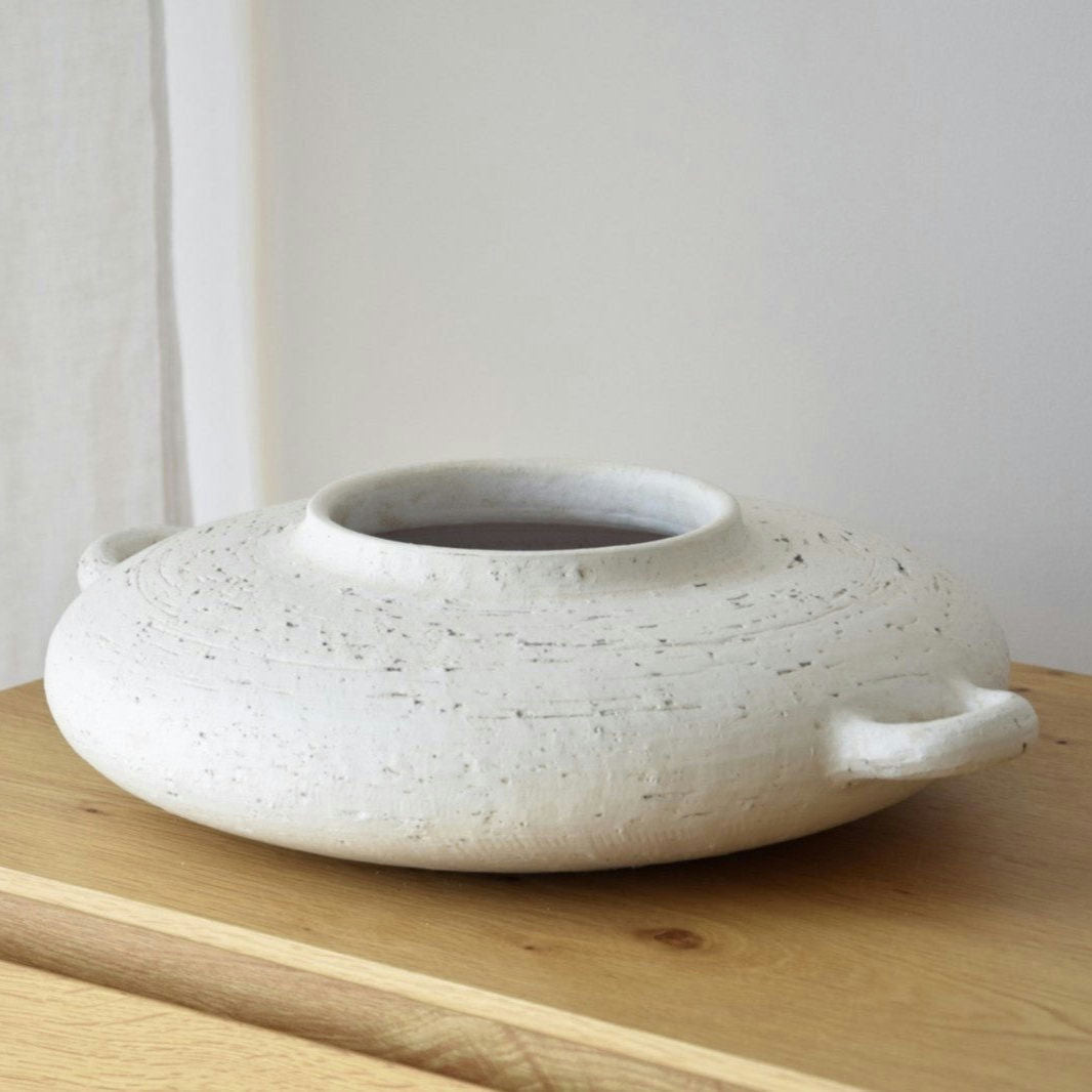 El Centro de Mesa Cerámica Ilda en Blanco Pátina, de forma redonda, acabado texturizado, amplia apertura y dos pequeñas asas, destaca sobre una superficie de madera.