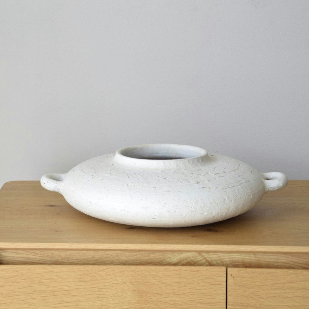 El Centro de Mesa Cerámica Ilda, Blanco Pátina, un jarrón redondo y plano con acabado blanco mate y dos asas laterales, adorna elegantemente una superficie de madera contra una pared lisa.