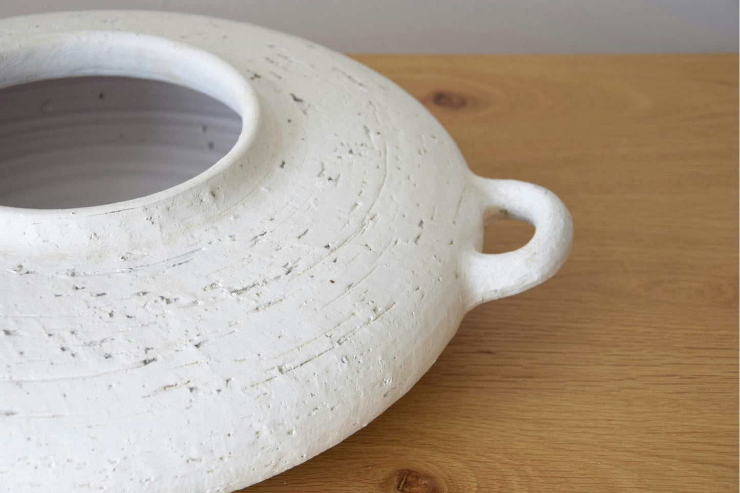 Primer plano del Centro de Mesa Cerámica Ilda, Blanco Pátina: un cuenco de cerámica blanca texturizada con asa sobre una superficie de madera. Su elegante acabado mate lo convierte en un centro de mesa artesanal ideal.