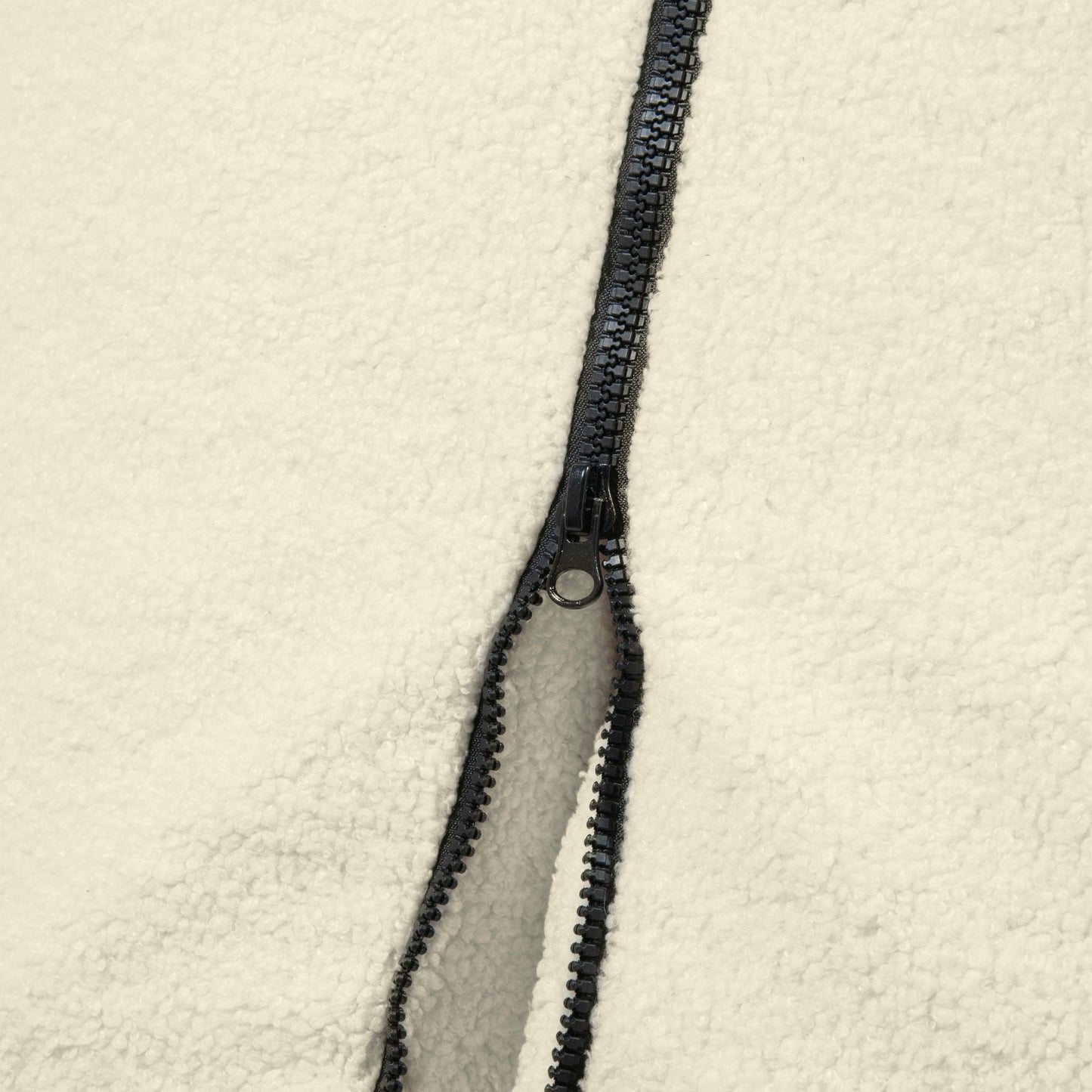 Primer plano de una cremallera negra parcialmente desabrochada sobre tejido polar blanquecino, que revela la acogedora textura del Bean bag puff grande de tela borreguito 115×90×75 cm - CLIP CLAP 115 TEDDY.