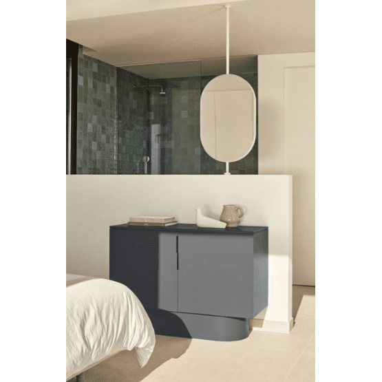 Un dormitorio minimalista con un armario gris oscuro de líneas elegantes, una cómoda de MDF con 2 Puertas Totem 2P, combinado con una colcha blanca, un espejo ovalado que refleja simplicidad y un área de ducha con azulejos, evoca una elegante estética minimalista.