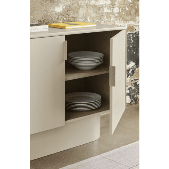 Una cómoda Totem 2P beige de MDF con 2 puertas y una puerta abierta muestra dos estantes con platos grises cuidadosamente apilados, lo que resalta el almacenamiento ordenado. Un libro sobre el gabinete realza su diseño moderno contra una pared parcialmente desgastada.