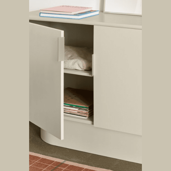 La Cómoda en MDF con 2 Puertas Totem 2P, en color beige, presenta un diseño moderno con una puerta abierta que muestra ropa doblada en el estante superior y libros debajo, ofreciendo un almacenamiento eficiente para cualquier habitación.
