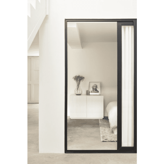 Un dormitorio minimalista cuenta con una cama blanca, la Cómoda en MDF con 2 Puertas y 2 Cajones Doric 2P2C para almacenamiento inteligente, una foto enmarcada y un jarrón con flores secas, todo elegantemente reflejado en un espejo de cuerpo entero con marco negro contra paredes blancas prístinas.