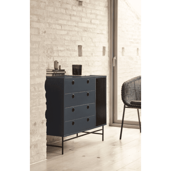 Una moderna escena de interior muestra la Cómoda en MDF con 4 Cajones y Puerta en Metal Punto 1P4C en azul oscuro, colocada contra una pared de ladrillos claros, acompañada por una silla de mimbre y una gran ventana. Esta disposición encarna el diseño industrial contemporáneo a la vez que ofrece soluciones de almacenamiento eficientes.