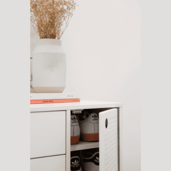 La cómoda de MDF con 4 cajones y puerta de metal Punto 1P4C, en un elegante color blanco, presenta una puerta ligeramente abierta que revela dos pares de zapatillas deportivas. Encima hay un jarrón blanco con flores secas y libros apilados. Sus puertas de metal perforado añaden un toque de almacenamiento moderno.