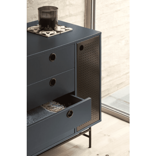 La cómoda de MDF con 4 cajones y puerta de metal Punto 1P4C, en azul oscuro, ofrece un estilo industrial contemporáneo. Un cajón está abierto con ropa cuidadosamente doblada y un objeto circular negro se encuentra sobre la cómoda junto a una gran ventana.