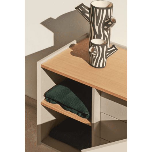 La Cómoda en MDF y Roble con 3 Cajones y Puerta Otto 1P3C rezuma diseño moderno con una manta verde y un elemento oscuro. Encima, un jarrón de cerámica con patrones en blanco y negro en forma de ramas de árbol agrega versatilidad y estilo al espacio.