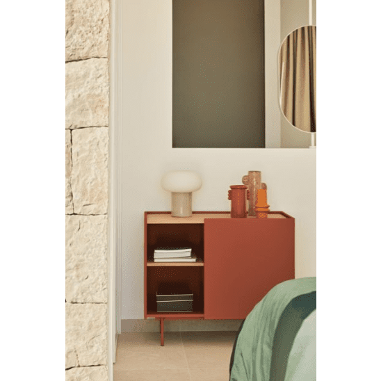 La Cómoda en MDF y Roble con 3 Cajones y Puerta Otto 1P3C, con diseño moderno, es un gabinete rojo compacto adornado con libros y decoración que incluye una lámpara y jarrones. Se apoya contra la pared junto a una cama con una manta verde, fusionando a la perfección versatilidad y estilo.