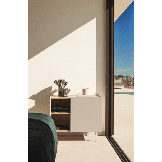Un dormitorio moderno presenta la Cómoda en MDF y Roble con 3 Cajones y Puerta Otto 1P3C, combinada con una mesita de noche blanca que sostiene un jarrón y una taza decorativos. Una gran ventana ofrece una vista soleada de una piscina al aire libre, lo que incorpora versatilidad y estilo en su diseño.