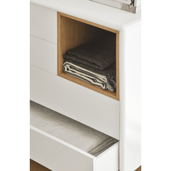 La Cómoda en MDF y Roble con 4 Cajones Corvo 4C captura el diseño moderno nórdico con un cajón abierto que muestra tela doblada y un estante abierto que contiene ropa cuidadosamente apilada, todo lo cual enfatiza el almacenamiento eficiente.