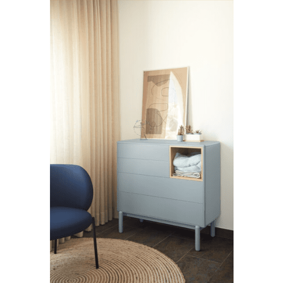 La Cómoda en MDF y Roble con 4 Cajones Corvo 4C en color gris, que muestra un estante abierto con toallas cuidadosamente dobladas, se encuentra contra una pared adornada con un estampado abstracto enmarcado. Cerca de allí, una silla azul reposa junto a cortinas beige sobre una alfombra tejida redonda, lo que realza el diseño nórdico moderno de la habitación.