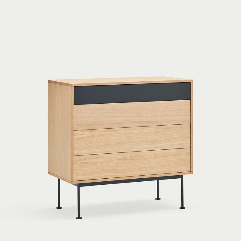 La Cómoda en MDF y Roble con cuatro cajones, incluido un cajón superior con un panel negro, se apoya sobre finas patas de metal negro. Incorpora un diseño minimalista sobre un fondo sencillo.