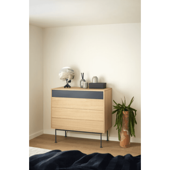 La Cómoda en MDF y Roble Yoko 4C con cubierta oscura se destaca contra una pared blanca, reflejando un diseño minimalista y ofreciendo un amplio espacio de almacenamiento contemporáneo. Está adornada con una planta a su lado y varios elementos decorativos en la parte superior, incluido un espejo.