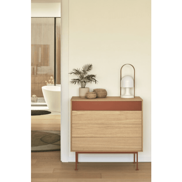La Cómoda en MDF y Roble con 4 Cajones Yoko 4C con tapa roja sostiene elegantemente una planta en maceta, una canasta y una lámpara, mostrando un diseño minimalista. Al fondo, a través de una puerta, se ve el baño con una bañera blanca.