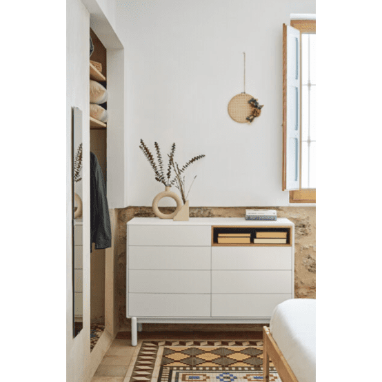 Un rincón minimalista del dormitorio presenta la Cómoda en MDF y Roble con 7 Cajones Corvo 7C, adornada con un jarrón decorativo y libros. Una ventana abierta, una decoración de pared y un piso de baldosas con motivos geométricos realzan el encanto del diseño interior.