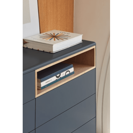 La Cómoda en MDF y Roble con 7 Cajones Corvo 7C es una cómoda azul con un estante de madera que sostiene un libro y una caja, mientras que su parte superior muestra un gran libro con motivos geométricos, añadiendo elegancia a cualquier diseño de interiores.