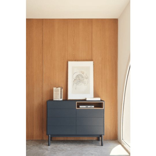 En una habitación minimalista, la Cómoda en MDF y Roble con 7 Cajones Corvo 7C sirve como tocador negro con un estante abierto que sostiene un libro y un marco de fotos contra una pared revestida de madera, resaltada por una pieza de arte abstracto que refleja un elegante diseño interior.