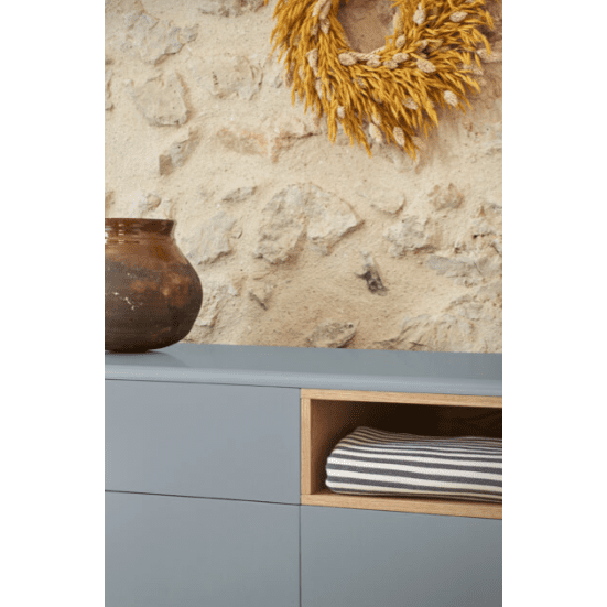 La cómoda de MDF y roble con 7 cajones Corvo 7C cuenta con un aparador azul con un compartimento de madera para tela de rayas doblada. Una maceta de cerámica marrón se encuentra encima y una corona de trigo decora la pared de piedra texturizada en el fondo, ideal para cualquier ambiente de diseño interior.