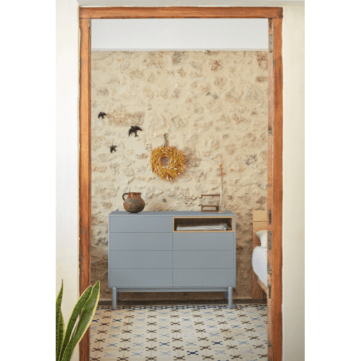 En una habitación acogedora, la cómoda en MDF y Roble con 7 Cajones Corvo 7C reposa contra una pared de piedra. La cómoda azul presenta una corona y pájaros negros, combinados con una maceta de cerámica sobre un piso de baldosas estampadas. Una planta en primer plano completa esta obra maestra del diseño de interiores.