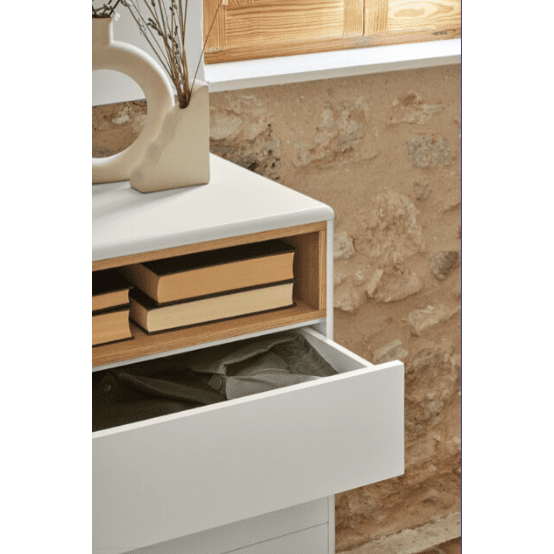 La Cómoda en MDF y Roble con 7 Cajones Corvo 7C se encuentra frente a una pared de piedra con una ventana de madera. Su fachada blanca muestra un cajón abierto lleno de tela, mientras que piezas de decoración como un jarrón y libros muestran un exquisito diseño interior en la parte superior.